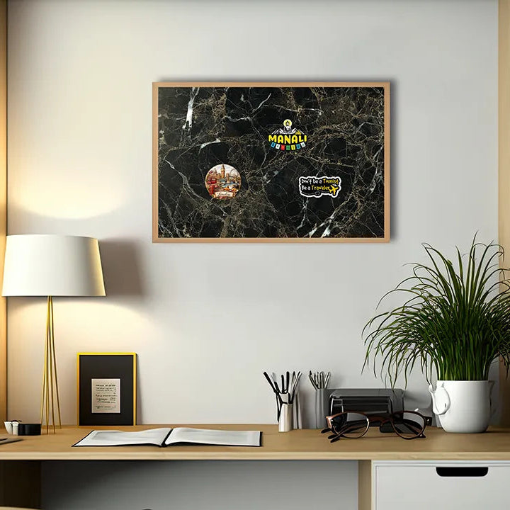 A framed black marble-style wall art print with colorful circular patches and the word 'MANGU' visible, hanging above a wooden desk in a modern home office setting with a lamp, computer, notebook, and potted plant.